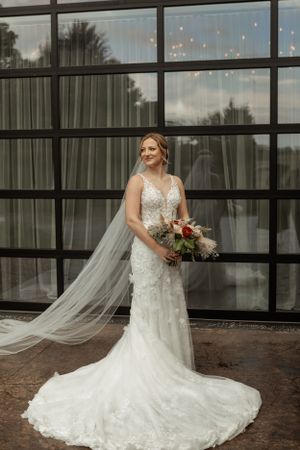 Bride in lace gown with cathedral veil poses against modern glass building.
