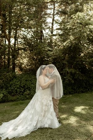 A romantic wedding moment captured in a sunlit garden with evergreen trees in the background.