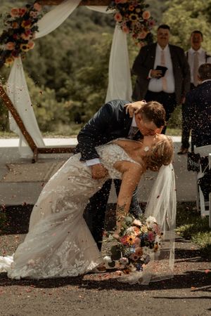 A romantic dip kiss under a floral wedding arch with rose petals falling around the couple.