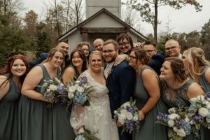 Wedding party in sage green dresses poses together outside rustic chapel.