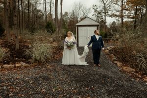 A couple in formal wedding attire walks together on a gravel path near a white garden shed on an autumn day.
