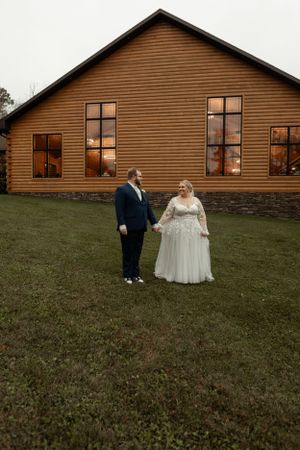 A couple stands together in formal attire against a rustic brown wooden venue building.