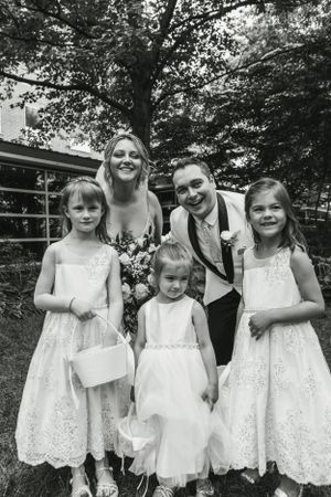 Wedding party portrait with flower girls in white lace dresses outdoors.