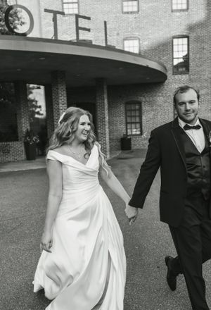 Black and white photo of couple running joyfully outside historic hotel entrance.