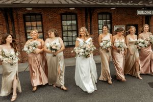 A wedding party poses together against a brick building exterior in coordinated champagne-colored attire.