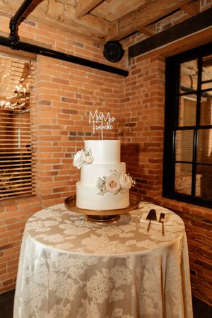 An elegant three-tier white wedding cake with floral decorations displayed against a brick wall.