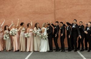 A wedding party poses playfully against a neutral wall in coordinated formal attire.