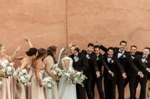 Wedding party in black tuxedos and blush dresses pose together in a candid moment with their bouquets.