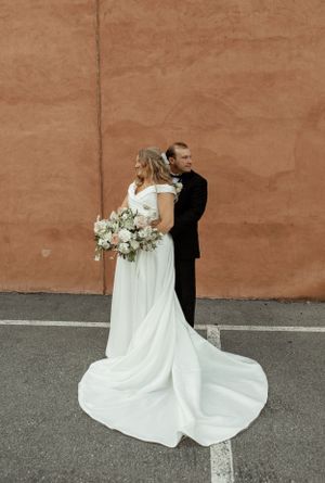 Newlywed couple poses against terra cotta wall with elegant white bouquet and flowing train.