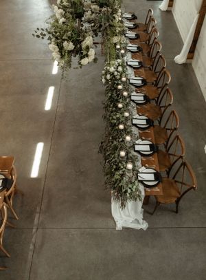 Overhead view of a wedding reception table with lush greenery runners and wooden cross-back chairs.