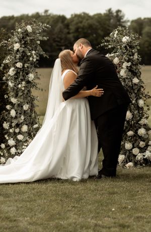 A romantic wedding ceremony moment under a white floral arch in an outdoor garden setting.