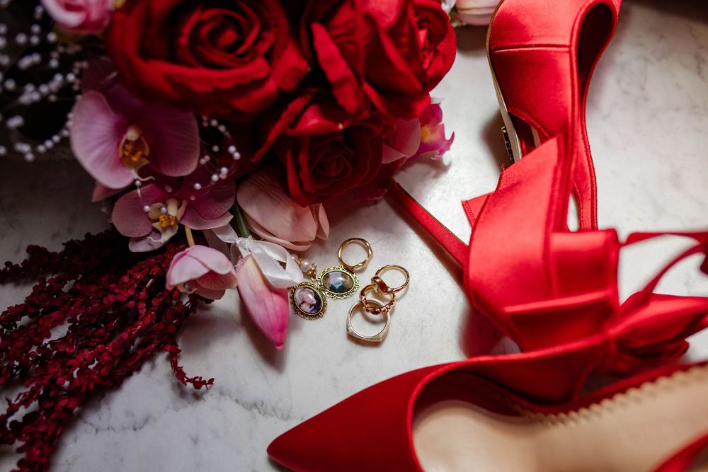 Red roses and jewelry arranged with elegant red high heel shoes on a marble surface.