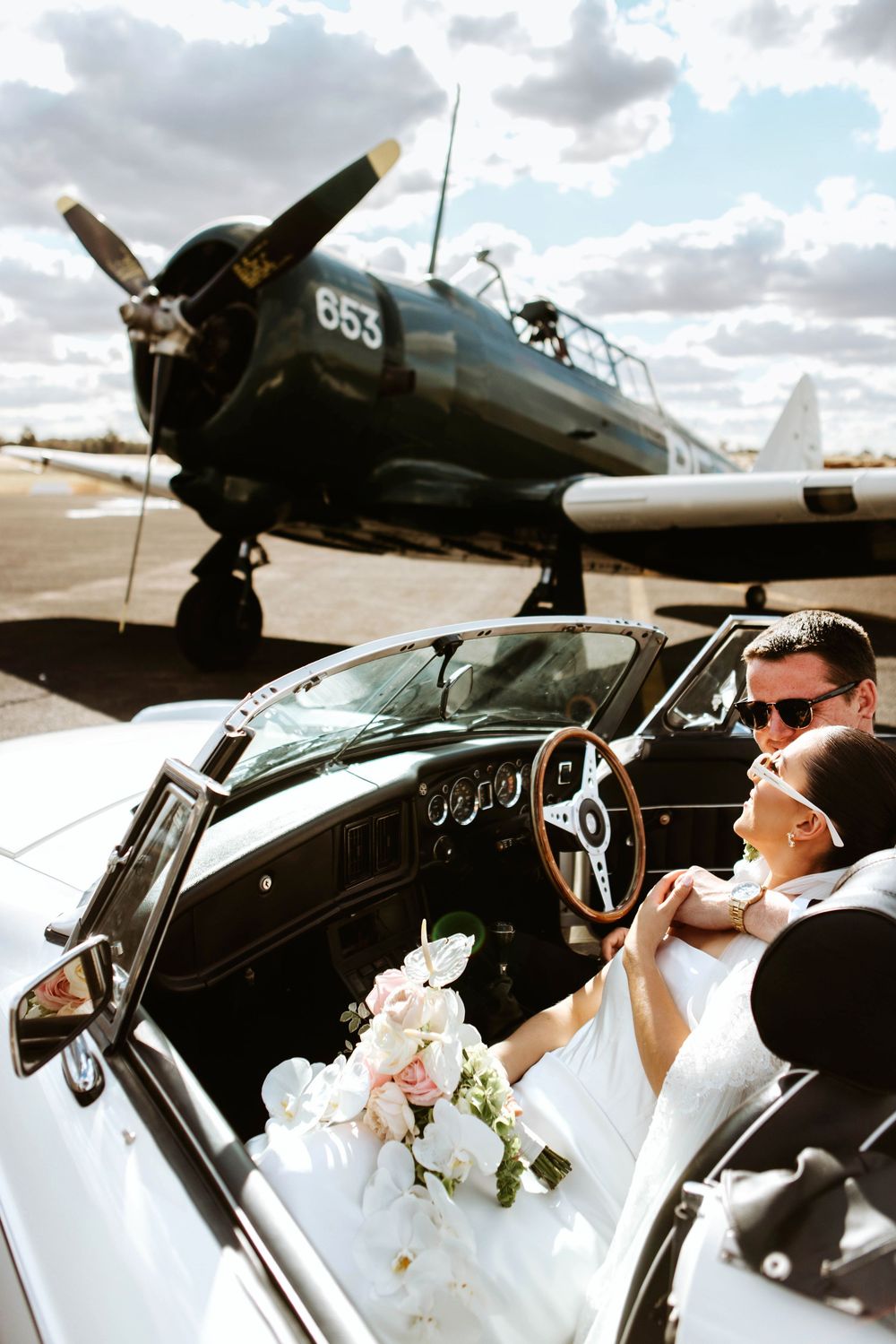Wedding couple in vintage convertible posing with historic military aircraft in background.