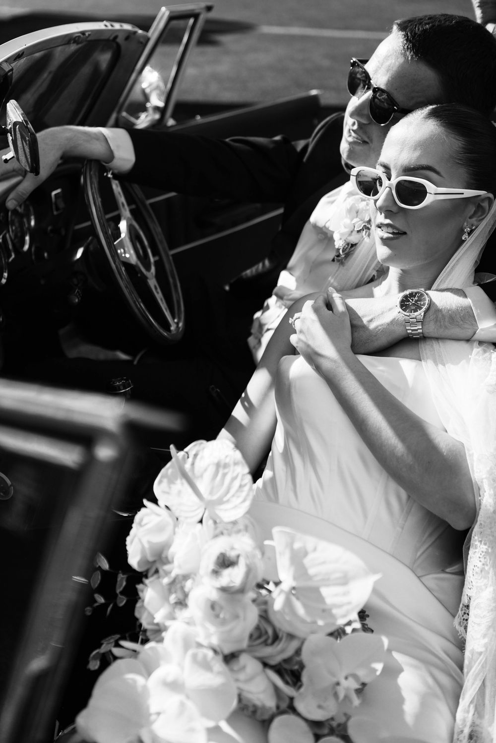 Vintage-style wedding photo of couple in classic car wearing retro sunglasses with white bouquet.