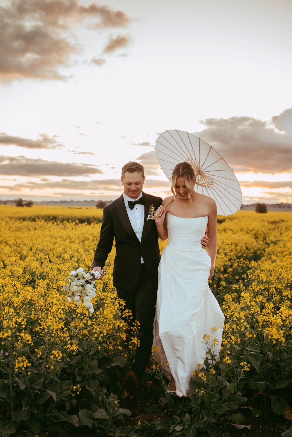 Couple with parasol walking through yellow canola field at sunset.