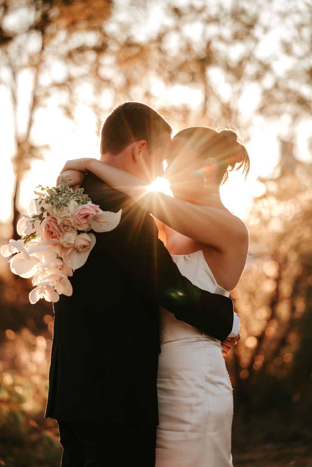 Romantic sunset wedding photo with couple embracing as sunlight streams through trees.