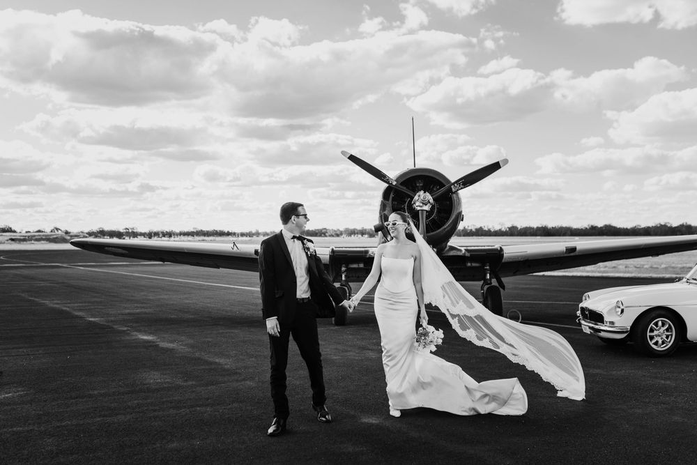 Elegant wedding couple posing with vintage airplane on runway in black and white.