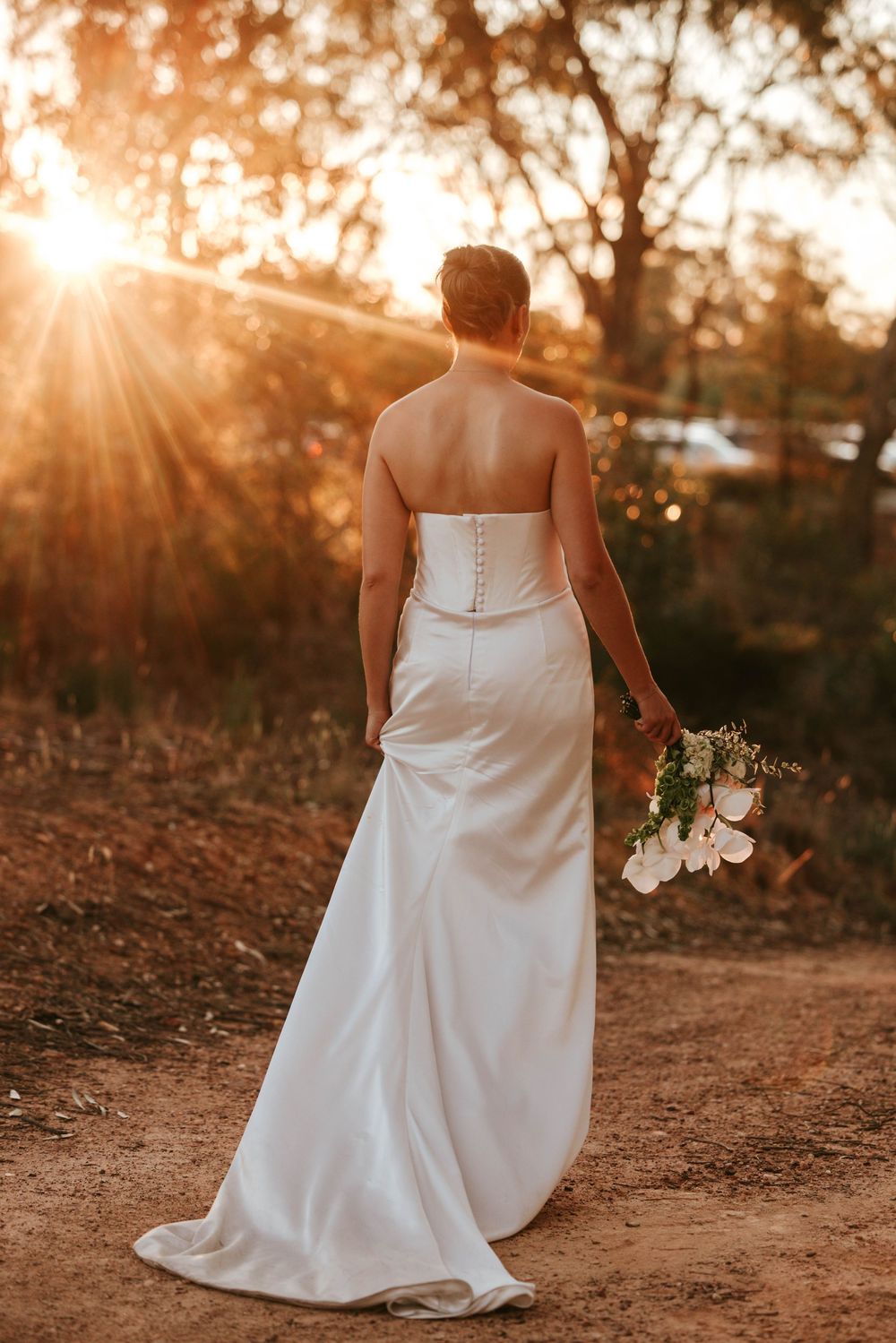 Bride in elegant white dress holding bouquet walks along sunset-lit path.