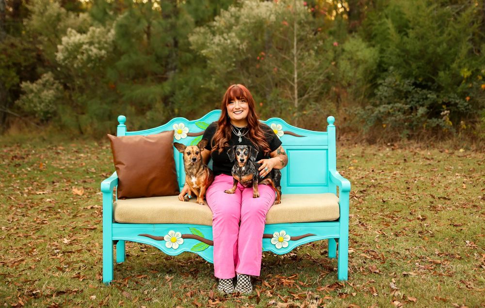 A woman sits on a turquoise floral bench outdoors with two small dogs on her lap.