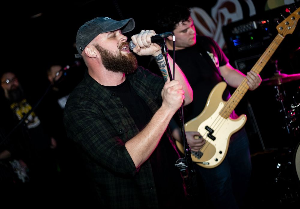 A vocalist performs on stage with a microphone while a bassist plays guitar behind him at a live concert.