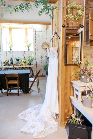 A delicate white lace wedding dress hangs in a rustic venue decorated with greenery and floral arrangements.
