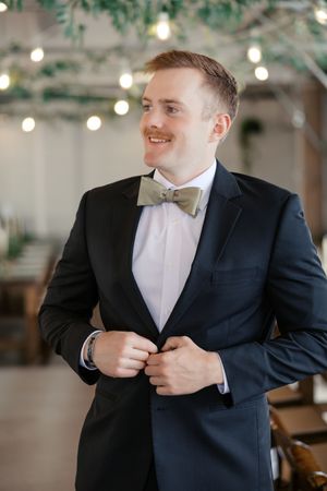 A groom in a black suit and bow tie adjusts his jacket while getting ready for a wedding ceremony.