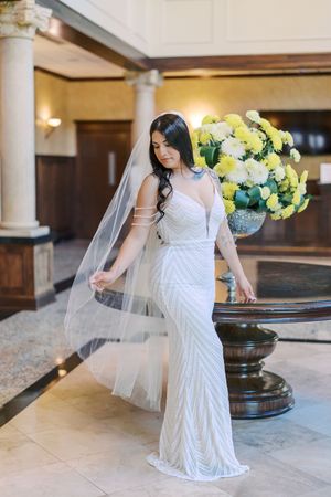A bride in a white lace gown and veil poses gracefully in an elegant venue with columns and floral arrangements.