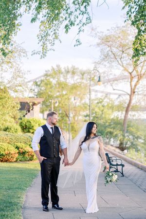A romantic couple in formal attire walks along a garden pathway during sunset, holding hands and smiling.