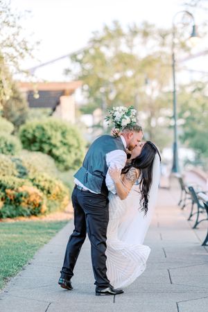 A romantic couple in wedding attire dips and kisses in a park setting with benches and greenery in the background.