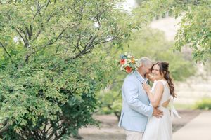 A couple shares a romantic kiss under lush green trees in an outdoor garden setting.