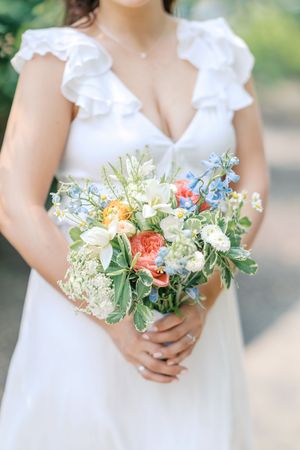 Delicate floral bouquets with coral roses, white flowers and greenery held against white dresses in natural light.