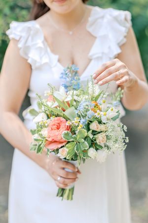 Delicate coral and white floral wedding bouquet with greenery held against a white dress in an outdoor garden setting.