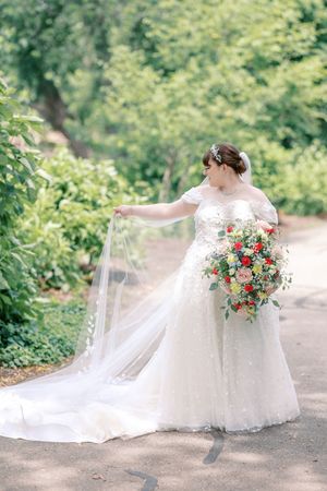 A bride in a flowing white wedding gown sits gracefully on a garden path holding a vibrant coral and white floral bouquet.