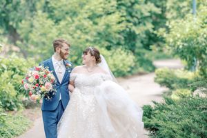 A couple in wedding attire walks along a garden path, with the bride holding a colorful bouquet and wearing a flowing white dress.