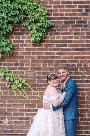 A couple poses against a brick wall with climbing ivy, wearing wedding attire on their special day.