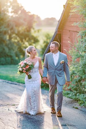 A romantic wedding couple walks together on a sunlit path beside a rustic red barn during golden hour.