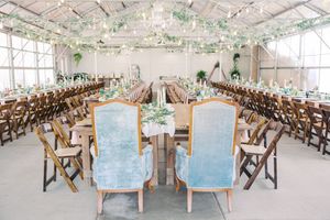 Elegant wedding reception setup with vintage blue chairs and wooden folding seats under a glass-enclosed conservatory space.