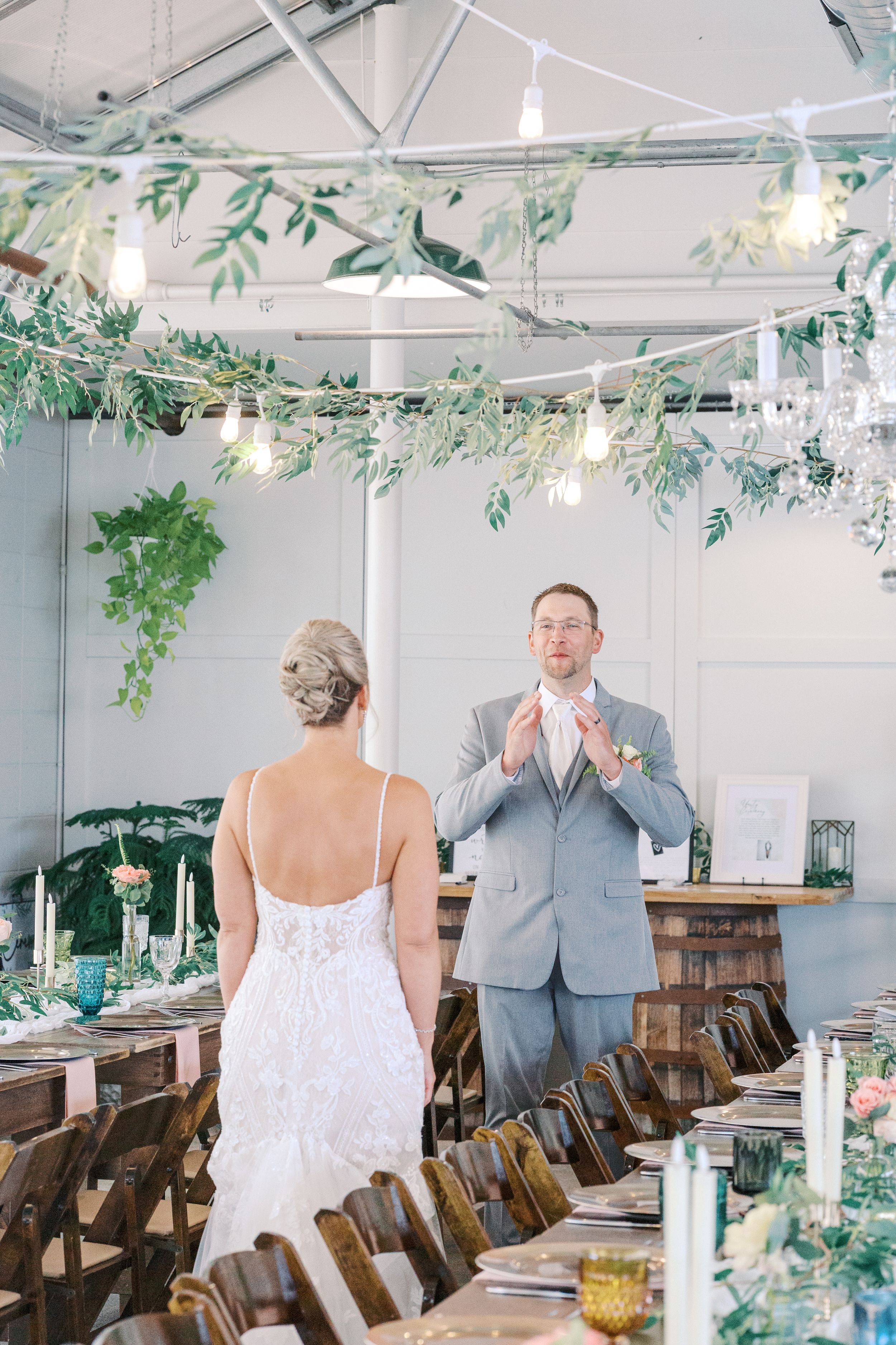 A wedding reception venue decorated with hanging greenery and lights as a couple shares a moment at the head table.
