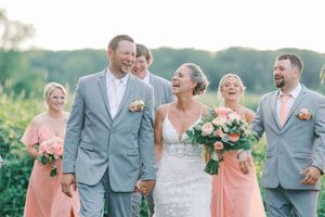 Wedding party in gray suits and coral dresses laughing together outdoors on a sunny day.