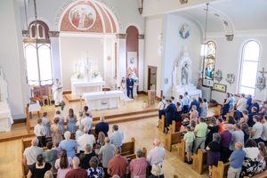 People gather inside a bright church sanctuary with white walls and arched windows for a religious service.