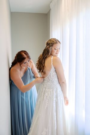 Bridesmaids in dusty blue dresses help button up an elegant lace wedding gown in a sunlit room.