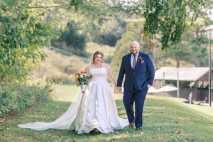 A couple walks hand in hand on a grassy hillside during their outdoor wedding ceremony on a sunny day.