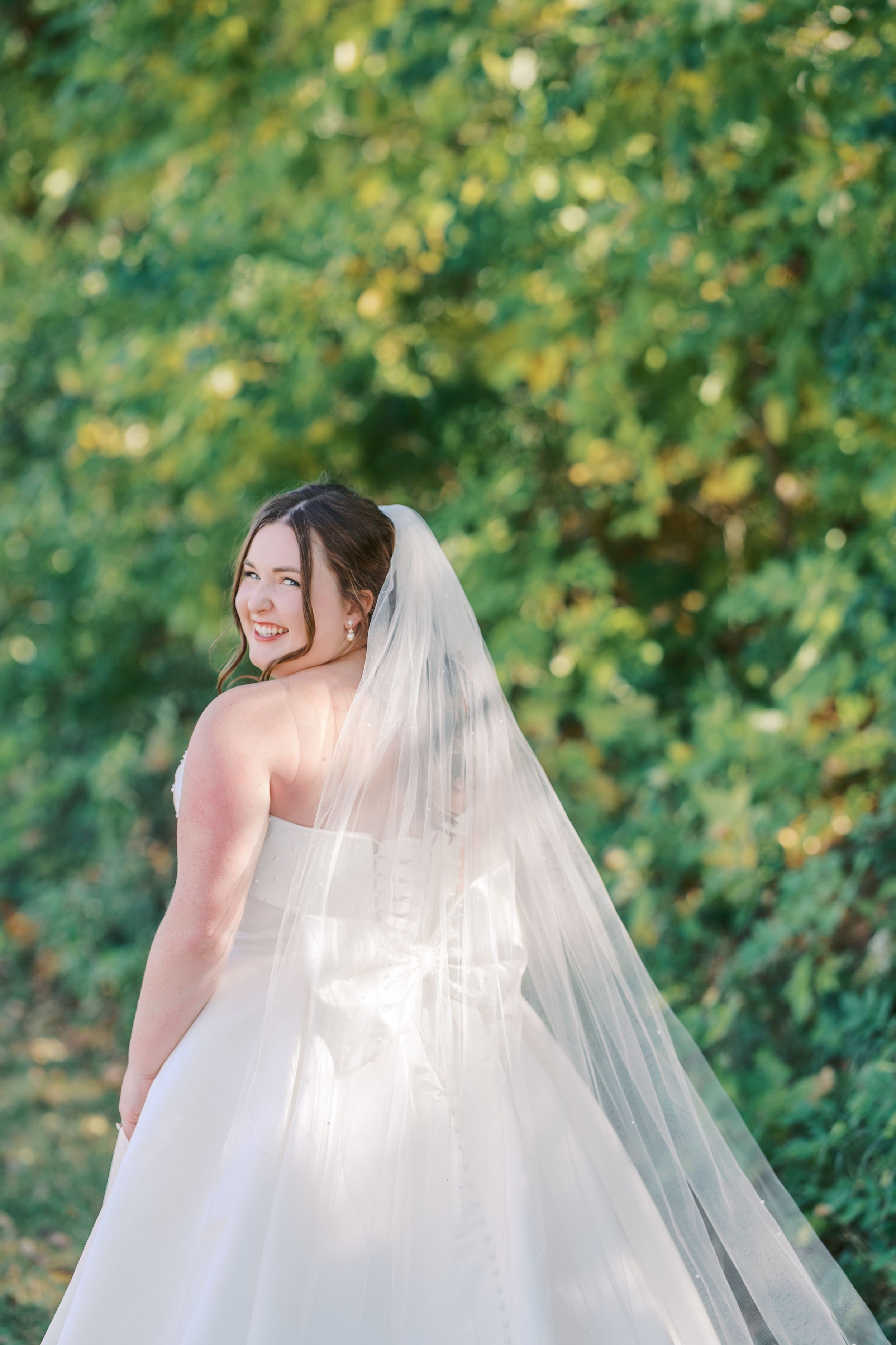 A bride in a flowing white wedding dress and veil poses outdoors against a soft green foliage background.