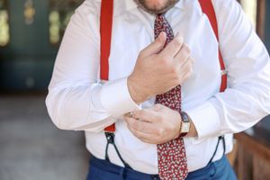 Professional wearing a white dress shirt and red suspenders adjusts a patterned necktie while standing indoors.
