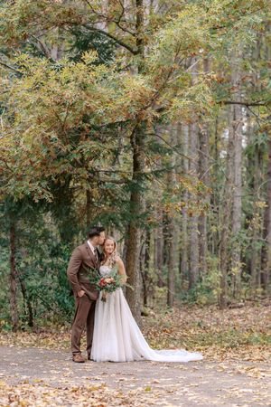 A romantic wedding couple shares a tender moment on a path covered in autumn leaves surrounded by trees.