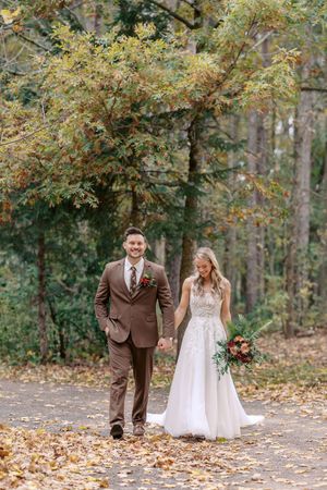 A couple in brown suit and white wedding dress walk hand in hand along a leaf-strewn forest path in autumn.