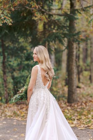 A person in a flowing white dress twirls gracefully in a wooded autumn setting with fallen leaves on the ground.