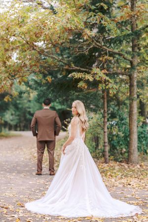 A couple in wedding attire walks through a fall forest path with autumn leaves scattered on the ground.