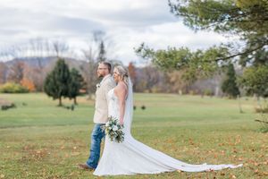 A couple stands in a scenic autumn field with fall foliage and rolling hills in the background.