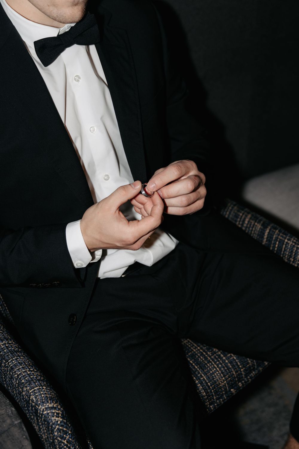 A person in formal attire adjusts their shirt cuff while wearing a black tuxedo and bow tie.